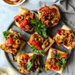 A rustic plate holds several pieces of roasted red pepper bruschetta, each topped with melted cheese, vibrant sweet peppers, olives, and fresh parsley. A drizzle of balsamic glaze adds a glossy finish. In the background, a soft blue napkin rests beside a small bowl of dark balsamic glaze with a spoon. A glass of white wine sits at the edge of the frame, complementing the dish's rich colors and textures.