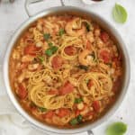 A stainless steel pan filled with cooked spaghetti, shrimp, white beans, diced tomatoes, and fresh basil leaves in a light tomato-based sauce. The pan is resting on a light-colored surface with a white cloth nearby. A small bowl of red pepper flakes and a few fresh basil leaves are placed next to the pan, along with stacked white plates and silver utensils in the top left corner.