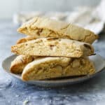 Four slices of biscotti (an Italian dessert biscuit) stacked on a plate set against a textured blue-gray background. The biscotti are golden brown and studded with nuts, creating a rustic and appetizing appearance. A blurred cloth is visible in the background.