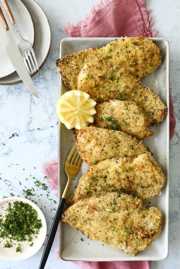 A rectangular plate with five pieces of breaded chicken garnished with chopped herbs. Theres a halved lemon slice on the side. A fork, knife, and two stacked plates are nearby. A small bowl of herbs and a pink cloth complete the arrangement.