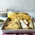 Slices of breaded and baked chicken breast on a tray, garnished with herbs. Lemon wedges are in the background. The tray rests on a pink cloth, with a soft-focus background suggesting a casual dining setting.