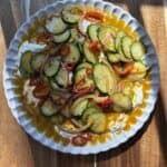 A white dish filled with a salad of sliced cucumbers, red onions, and red chili peppers, drizzled with a tangy dressing, sits on a wooden surface with sunlight highlighting its vibrant colors.