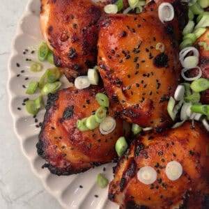 Plate of glazed, grilled chicken thighs garnished with chopped green onions and black sesame seeds, served on a scalloped white dish on a marble surface.