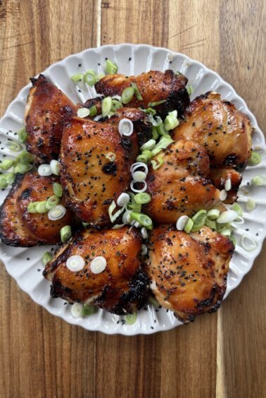 A plate of glazed, roasted miso chicken thighs garnished with sliced green onions, served on a white scalloped dish atop a wooden surface.