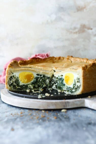 A pie with a golden crust, sliced to reveal layers of whole eggs and spinach filling inside, displayed on a round cutting board.