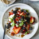 A white plate with a fresh berries and burrata salad of strawberries, blueberries, blackberries, cherries, basil leaves, and dollops of cream, drizzled with balsamic glaze. A fork and a bowl of dressing sit nearby on a gray surface.