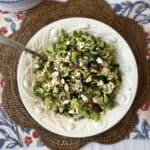 A white plate holds a broccoli salad with chopped vegetables, seeds, and crumbled cheese, placed on a woven placemat atop a decorated tablecloth. A serving spoon rests in the bowl, and stacked plates are nearby.