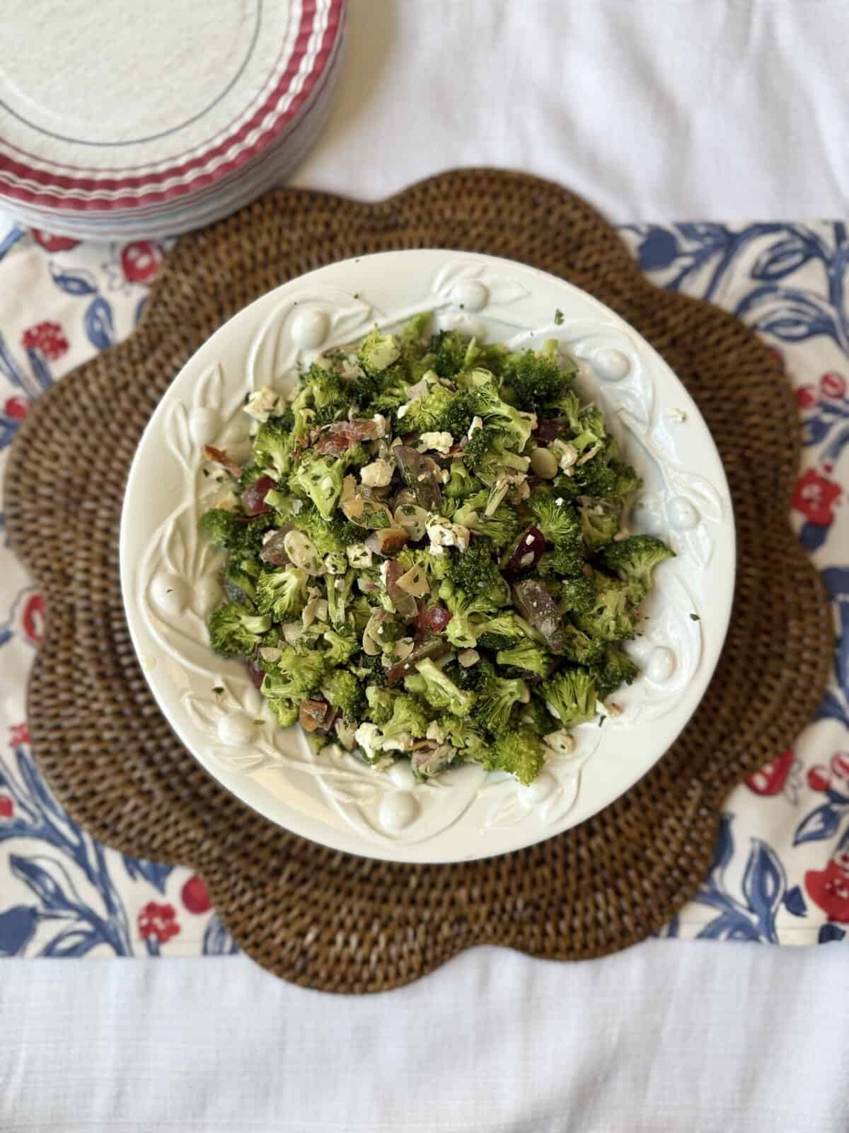 A white bowl filled with broccoli salad containing shredded cheese and chopped vegetables sits on a woven placemat over a floral tablecloth, with stacked plates nearby.
