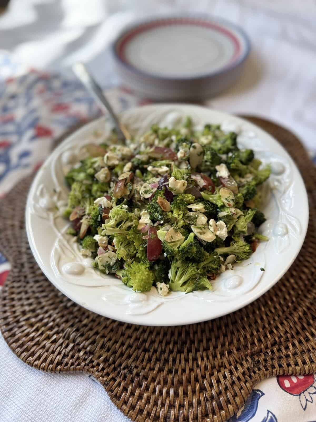 A white platter of broccoli salad with sliced almonds, feta cheese, and red grapes sits on a woven placemat. A serving spoon rests in the salad, and a stack of plates is in the background on a patterned tablecloth.