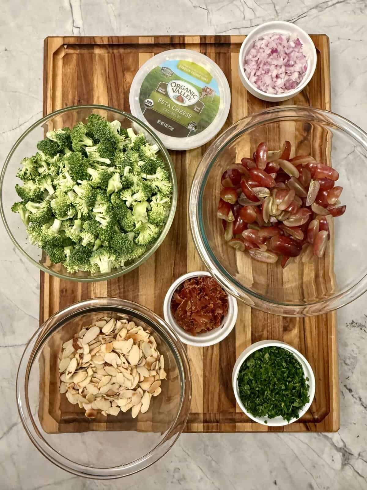 A wooden board holds bowls of broccoli florets, sliced red grapes, slivered almonds, chopped red onion, crumbled goat cheese, chopped parsley, and crispy bacon bits, all arranged neatly on a marble countertop.