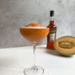 A frozen Aperol spritz in a cocktail glass filled with an orange frozen cantaloup slushy is on a white surface. In the background, there is a bottle of Aperol and a halved cantaloupe melon. The setting is minimal with a neutral backdrop.