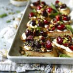 A tray of toasted bread slices topped with goat cheese, fresh cherries, chopped pistachios, and herbs, drizzled with balsamic glaze, sits on a light blue surface with a bowl of extra pistachios in the background.