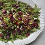 A platter of fresh arugula salad topped with halved cherries, crumbled cheese, chopped pistachios, and sliced red chili on a white plate with blue stripes.