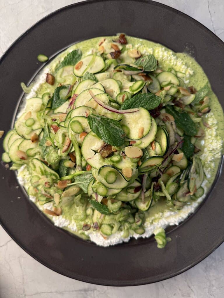 A fresh zucchini salad with thinly sliced zucchini, mint leaves, slivered almonds, and red onions on a creamy green herb sauce, served on a dark plate.