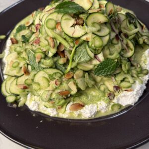 A fresh salad with thinly sliced zucchini, pine nuts, herbs, and nuts on a bed of creamy ricotta cheese, served on a dark plate atop a light marble surface.