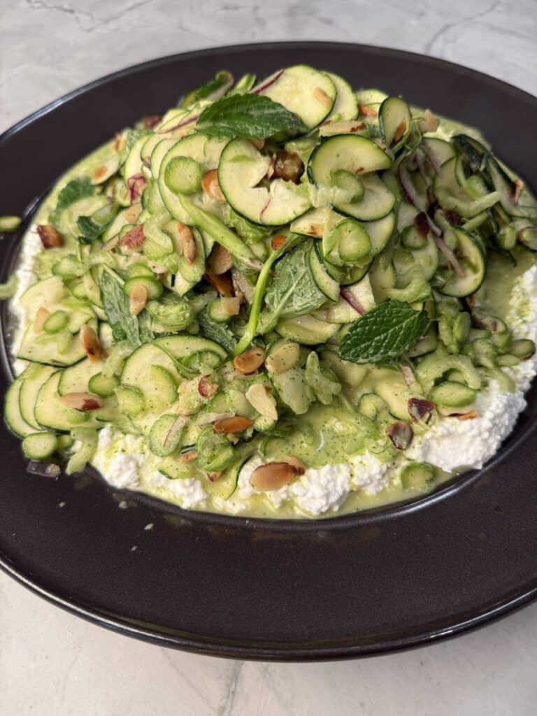 A raw detox salad with thinly sliced zucchini, pine nuts, herbs, and nuts on a bed of creamy ricotta cheese, served on a dark plate atop a light marble surface.