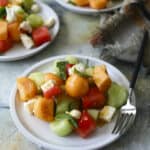 Three plates of fresh salad with cubes of watermelon, cantaloupe, honeydew melon, cucumber, and feta cheese are on a rustic surface. A fork and folded napkin rest beside one plate. Mint leaves garnish the salads.
