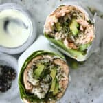 Two halves of a sushi burrito filled with rice, avocado, greens, and fish are placed on a round plate, with a small bowl of creamy dipping sauce and another bowl with dark seasoning on the side.