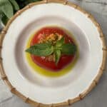A stuffed tomato topped with fresh basil sits in olive oil on a white plate with a bamboo-patterned rim; a bunch of basil leaves and a patterned cup are nearby on a marble surface.
