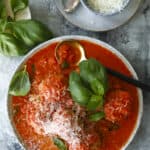 A bowl of meatballs in tomato sauce garnished with fresh basil and shredded Parmesan cheese, with a spoon and a small bowl of extra grated cheese beside the plate.