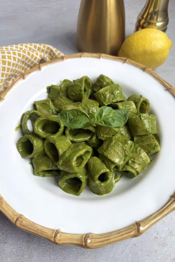 A white plate with short, tube-shaped pasta coated in green pesto sauce, garnished with fresh basil leaves. A yellow lemon, brass containers, and a yellow-patterned napkin are in the background.