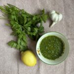 A bowl of green sauce called gremolata sits on a beige cloth next to fresh parsley, a lemon, and a bulb of garlic.