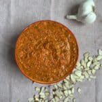 A red bowl filled with a chunky red Romesco sauce or dip sits on a beige cloth, next to a whole garlic bulb and scattered sliced almonds.
