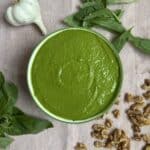 A bowl of green pesto sauce on a cloth, surrounded by fresh basil leaves, a head of garlic, and scattered walnut pieces.