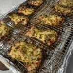 Cooked, marinated chicken pieces rest on a wire rack over a foil-lined baking tray. The chicken is golden-brown with charred edges and visible herbs. The tray sits on a light marble surface.