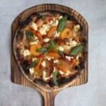 A rustic peach pizza on a wooden paddle topped with fresh basil, sliced peaches, red onion, and dollops of cheese, set against a light textured background.