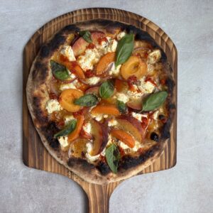 A rustic peach pizza on a wooden paddle topped with fresh basil, sliced peaches, red onion, and dollops of cheese, set against a light textured background.