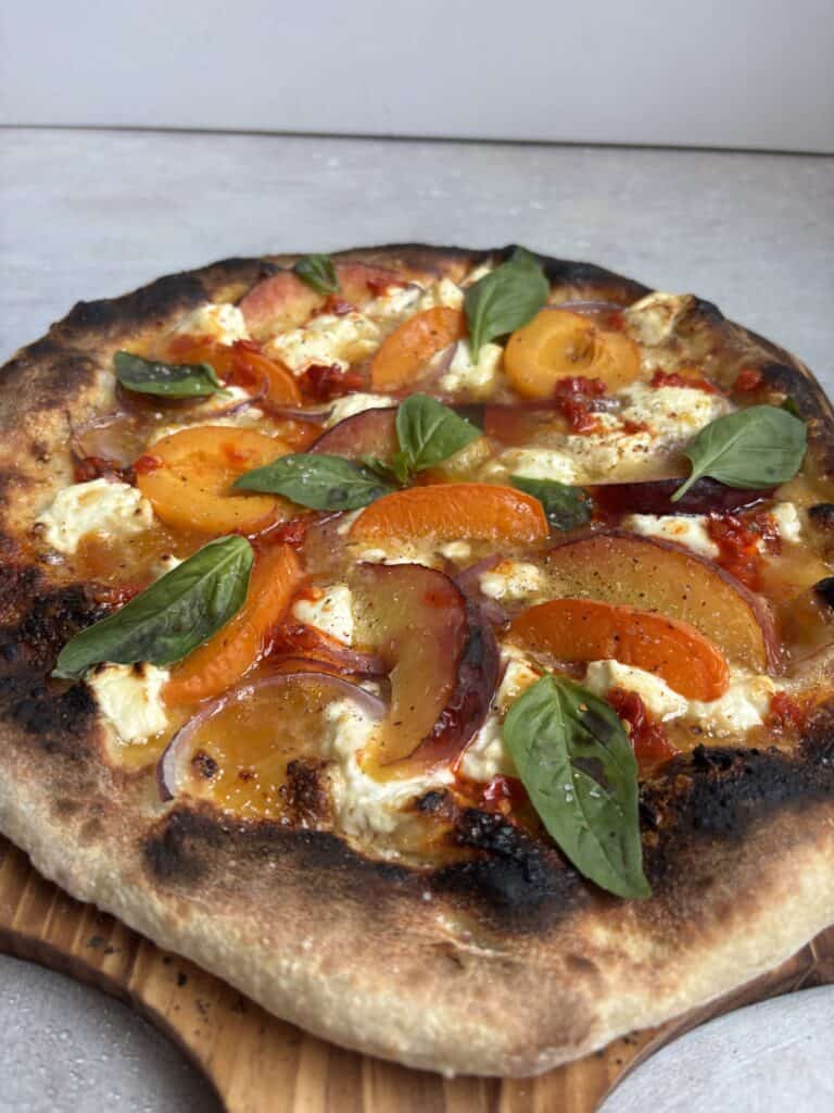 A freshly baked pizza topped with sliced yellow and orange heirloom tomatoes, dollops of ricotta cheese, red onion, fresh basil leaves, and a crispy, slightly charred crust on a wooden board.