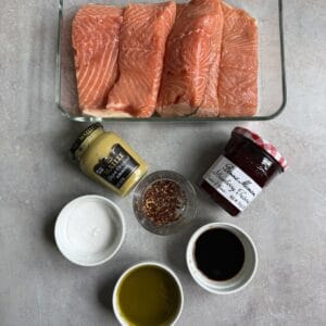 Four raw salmon fillets in a glass dish, with small bowls of olive oil, salt, red pepper flakes, soy sauce, Dijon mustard, and a jar of Bonne Maman raspberry preserves arranged below.