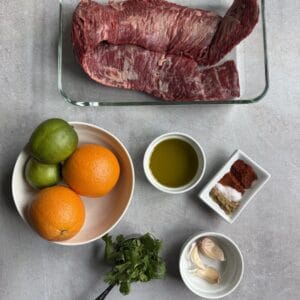 Meal prep raw skirt steak in a glass dish, surrounded by oranges, limes, olive oil, spices, fresh cilantro, and garlic cloves, arranged neatly on a light gray surface.