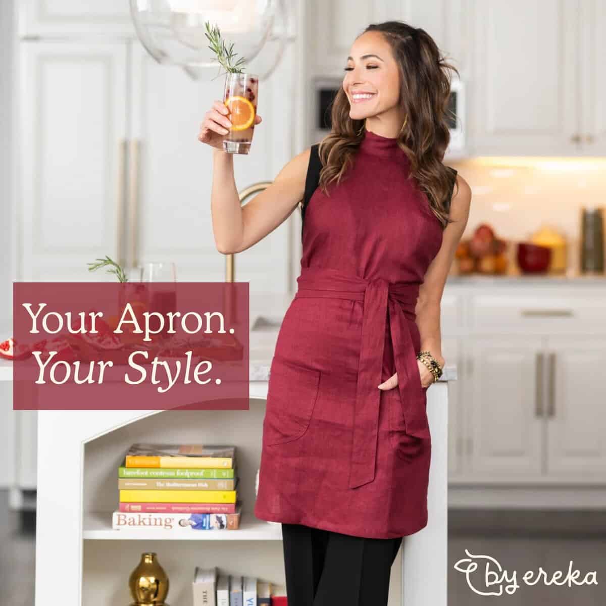 A smiling woman in a modern kitchen wears a stylish maroon apron, holding a drink garnished with herbs. Text reads Your Apron. Your Style. The Byereka logo is in the lower right corner.