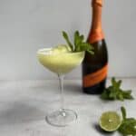 A frozen cocktail garnished with fresh mint is served in a coupe glass. Behind it, there is a bottle of sparkling wine, a halved lime, and more mint leaves on a light surface.