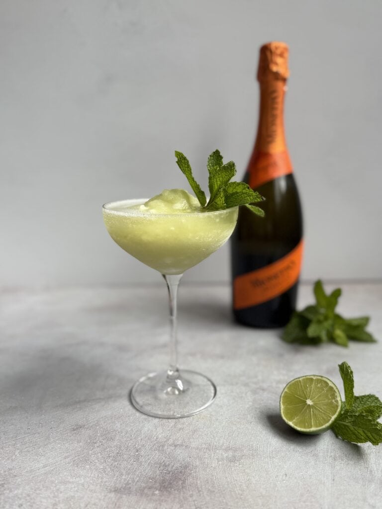 A frozen cocktail garnished with fresh mint is served in a coupe glass. Behind it, there is a bottle of sparkling wine, a halved lime, and more mint leaves on a light surface.