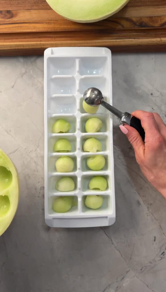 A hand uses a melon baller to place round honeydew melon balls into an ice cube tray on a gray countertop, with a half-cut melon beside the tray.