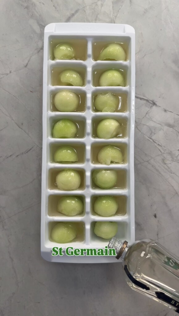 An ice cube tray filled with round green melon balls and liquid is set on a marble surface. A hand is pouring clear St Germain liqueur into one of the compartments.