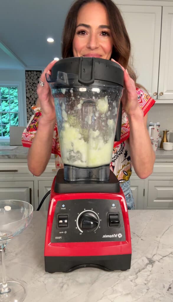 A woman in a colorful top holds the lid of a red Vitamix blender, blending a creamy mixture in a bright kitchen with white cabinets and a window in the background.