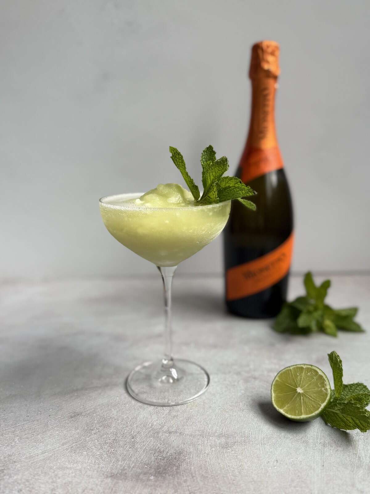 A coupe glass filled with a frozen Hugo Spritz, a pale green cocktail garnished with mint, sits in front of an orange-labeled sparkling wine bottle. There are mint sprigs and a halved lime on the surface beside the glass.