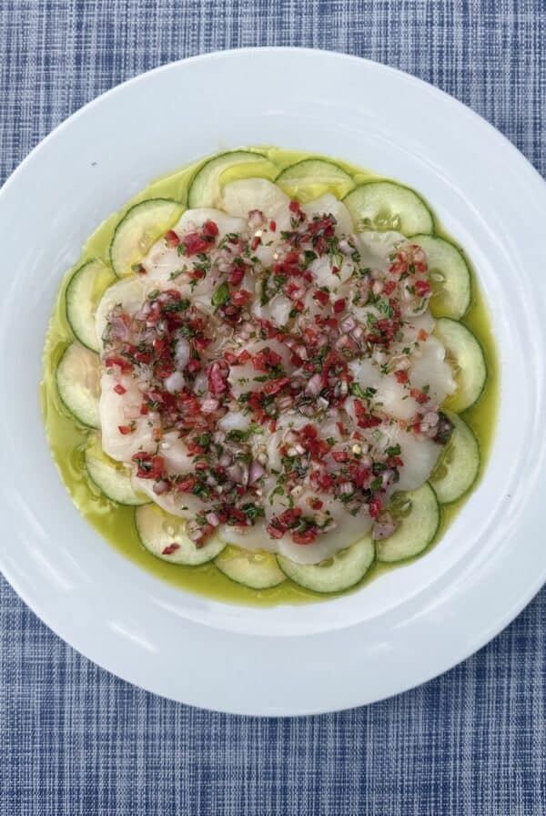 A white plate with thinly sliced cucumbers arranged in a circle, topped with a layer of sliced scallops and a colorful mixture of diced onions, herbs, and peppers, drizzled with olive oil, on a blue woven tablecloth.