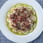 A white plate with thin cucumber slices arranged in a circle, topped with scallop crudo, finely diced red onion, herbs, and a drizzle of olive oil, placed on a blue textured tablecloth.