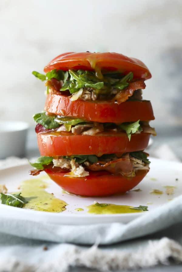 A stack of thick tomato slices layered with leafy greens and shredded chicken on a white plate, drizzled with olive oil, creating a fresh and colorful BLT Tomato Stack.