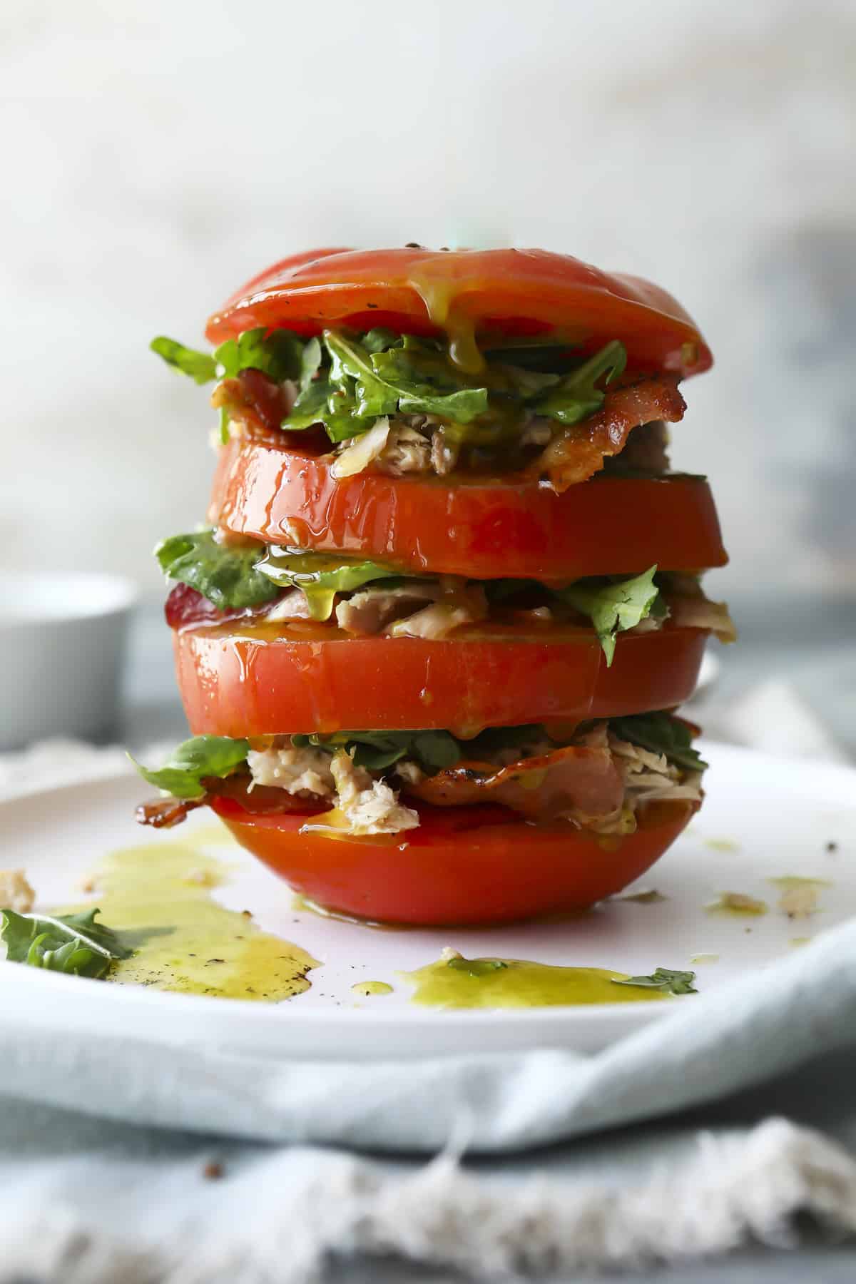 A stack of thick tomato slices layered with leafy greens and shredded chicken on a white plate, drizzled with olive oil, creating a fresh and colorful salad tower.