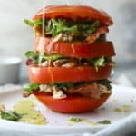 A stack of thick tomato slices layered with greens and shredded chicken, drizzled with olive oil, sits on a white plate.