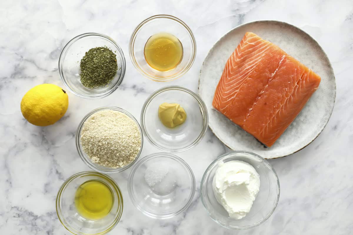 A plate with a raw salmon fillet next to small glass bowls containing breadcrumbs, olive oil, dried herbs, honey, Dijon mustard, yogurt, salt, and a whole lemon, all arranged on a marble surface.