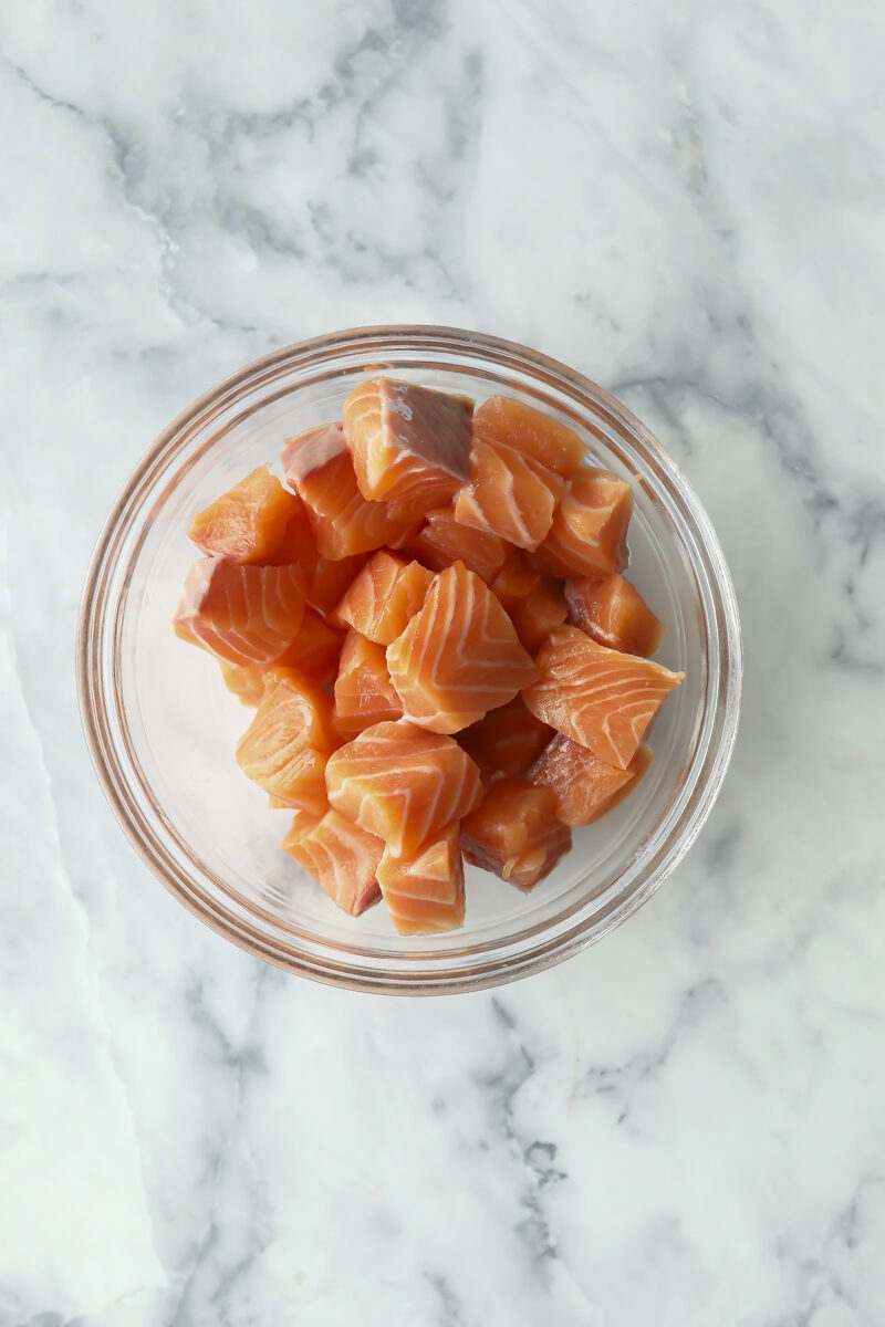 A glass bowl filled with cubed raw salmon sits on a white marble surface.