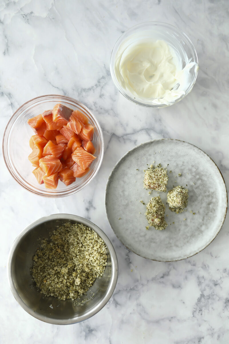 Four bowls and plates on a marble surface: minced herbs or breadcrumbs in a metal bowl, raw salmon pieces in a glass bowl, creamy white cheese or yogurt in a glass bowl, and two coated pieces on a gray plate.
