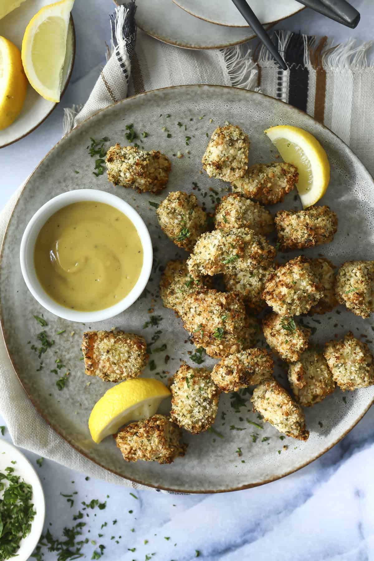 A plate of baked salmon bites garnished with chopped herbs, served with lemon wedges and a small bowl of creamy dipping sauce on a gray plate.